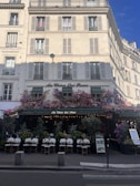 A charming Parisian café with a classic exterior, featuring an abundance of flowers draped over the entrance and windows. The seating area is set up with elegant white chairs and small tables, inviting passersby. The building has a cream facade with traditional French windows and shutters.