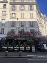 A charming Parisian café with a classic exterior, featuring an abundance of flowers draped over the entrance and windows. The seating area is set up with elegant white chairs and small tables, inviting passersby. The building has a cream facade with traditional French windows and shutters.