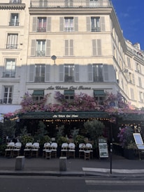 A charming Parisian café with a classic exterior, featuring an abundance of flowers draped over the entrance and windows. The seating area is set up with elegant white chairs and small tables, inviting passersby. The building has a cream facade with traditional French windows and shutters.