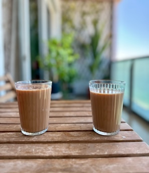 Two glasses filled with a brown liquid, possibly tea or coffee, are placed on a wooden table. The table is on a balcony or outdoor setting with a blurred background showing plants and a railing. The environment appears relaxed and serene.