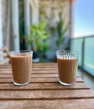 Two glasses filled with a brown liquid, possibly tea or coffee, are placed on a wooden table. The table is on a balcony or outdoor setting with a blurred background showing plants and a railing. The environment appears relaxed and serene.