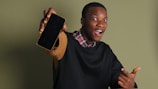 A person wearing a black sweater over a checkered shirt is holding a smartphone with enthusiasm. The background is plain green, and the individual displays an expression of excitement.