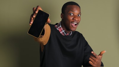 A person wearing a black sweater over a checkered shirt is holding a smartphone with enthusiasm. The background is plain green, and the individual displays an expression of excitement.