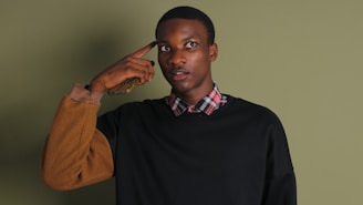 A person is standing with one hand holding a phone to their ear, wearing a plaid shirt under a black sweater with brown sleeves. The background is a solid olive green. The person appears to be looking upwards.