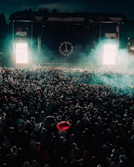 A large crowd gathered in front of a stage with illuminated signage and a peace symbol. The scene is filled with dense fog and a distant spotlight casts an ethereal glow over the crowd. A striking red umbrella stands out among the many people.