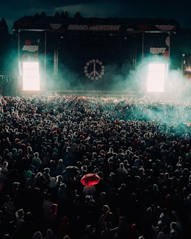 A large crowd gathered in front of a stage with illuminated signage and a peace symbol. The scene is filled with dense fog and a distant spotlight casts an ethereal glow over the crowd. A striking red umbrella stands out among the many people.