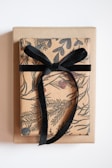 A neatly wrapped gift box with brown paper featuring black botanical illustrations and adorned with a black velvet ribbon tied in a bow. The gift is placed on a plain brown surface or another box, against a white background.
