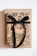 A neatly wrapped gift box with brown paper featuring black botanical illustrations and adorned with a black velvet ribbon tied in a bow. The gift is placed on a plain brown surface or another box, against a white background.