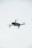 A quadcopter drone is hovering in the air against a plain, light-colored background. The drone has a compact design with four propellers.