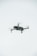 A quadcopter drone is hovering in the air against a plain, light-colored background. The drone has a compact design with four propellers.