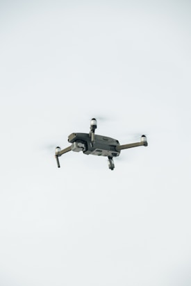 A quadcopter drone is hovering in the air against a plain, light-colored background. The drone has a compact design with four propellers.