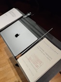 A music stand holds a closed laptop with an apple logo, a baton, and sheet music titled 'Troisième Symphonie' by Pierre Wissmer. The stand is set on a wooden floor.