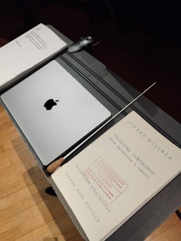 A music stand holds a closed laptop with an apple logo, a baton, and sheet music titled 'Troisième Symphonie' by Pierre Wissmer. The stand is set on a wooden floor.