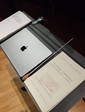 A music stand holds a closed laptop with an apple logo, a baton, and sheet music titled 'Troisième Symphonie' by Pierre Wissmer. The stand is set on a wooden floor.