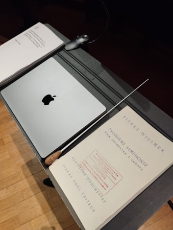 A music stand holds a closed laptop with an apple logo, a baton, and sheet music titled 'Troisi&egrave;me Symphonie' by Pierre Wissmer. The stand is set on a wooden floor.