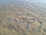 The word 'HAPPY' is written in large, capital letters on a sandy beach, accompanied by a simple, smiling face drawn above it. Footprints and small wave patterns are visible in the sand, along with a few small clumps of seaweed. The sand appears slightly damp, with the tide gently approaching in the background.