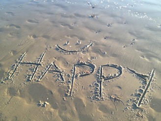 The word 'HAPPY' is written in large, capital letters on a sandy beach, accompanied by a simple, smiling face drawn above it. Footprints and small wave patterns are visible in the sand, along with a few small clumps of seaweed. The sand appears slightly damp, with the tide gently approaching in the background.