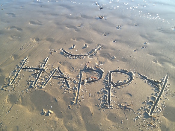 The word 'HAPPY' is written in large, capital letters on a sandy beach, accompanied by a simple, smiling face drawn above it. Footprints and small wave patterns are visible in the sand, along with a few small clumps of seaweed. The sand appears slightly damp, with the tide gently approaching in the background.
