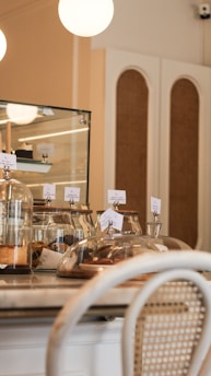 A cozy café interior with a countertop displaying assorted pastries under glass domes. Warm lighting and a mix of wooden and wicker furniture create a welcoming atmosphere.