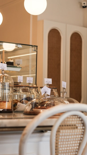 A cozy caf&eacute; interior with a countertop displaying assorted pastries under glass domes. Warm lighting and a mix of wooden and wicker furniture create a welcoming atmosphere.