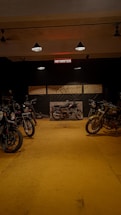 A dimly lit motorcycle garage featuring several motorcycles on a yellow floor. Various tools are displayed on the walls, and an illuminated sign reads 'MOTORIFICS'. The atmosphere is rustic and industrial.
