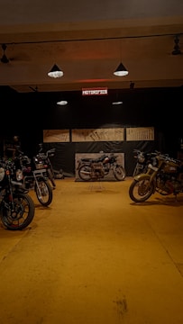 A dimly lit motorcycle garage featuring several motorcycles on a yellow floor. Various tools are displayed on the walls, and an illuminated sign reads 'MOTORIFICS'. The atmosphere is rustic and industrial.