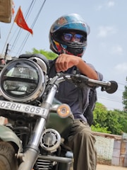 A person wearing a helmet and reflective sunglasses is seated on a large motorcycle. The motorcycle has a visible front headlight and a registration plate. The person is wearing a backpack and the background includes a red flag, wires, and some greenery.