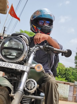A person wearing a helmet and reflective sunglasses is seated on a large motorcycle. The motorcycle has a visible front headlight and a registration plate. The person is wearing a backpack and the background includes a red flag, wires, and some greenery.