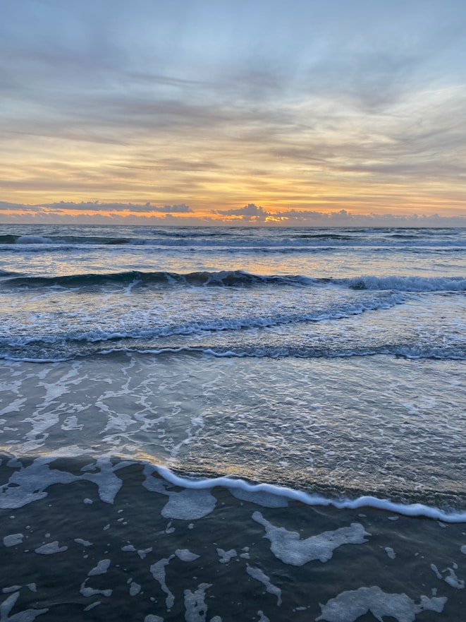 A serene view of the Maui coastline at sunrise, with gentle waves lapping against the shore near the beachfront rentals.