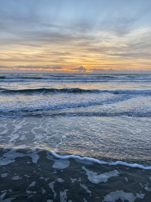 A serene beach scene at sunrise with gentle waves lapping against the shore. The sky is filled with soft clouds, colored in pastel shades by the emerging sun.