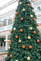 A large, decorated Christmas tree inside a commercial building. It is adorned with orange and gold baubles, with small white lights evenly distributed throughout its green branches. The surrounding environment has tall glass windows and ornate architectural details, creating a festive indoor atmosphere.