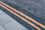 A close-up view of a road with two parallel yellow paint lines running along the dark asphalt. The texture of the road surface is rough and appears to be made of small gravel pieces. There is a section of light gray pavement to the left, made up of rectangular tiles.