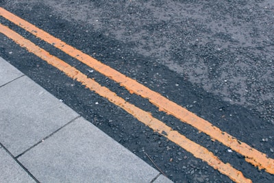 A close-up view of a road with two parallel yellow paint lines running along the dark asphalt. The texture of the road surface is rough and appears to be made of small gravel pieces. There is a section of light gray pavement to the left, made up of rectangular tiles.