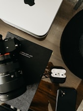 A workspace setup featuring a camera with a large lens, an open case containing wireless earbuds, a smartphone, a notebook titled 'Minimalism Art,' and a Mac computer. The items are arranged on a wooden surface, projecting a sleek and professional aesthetic.