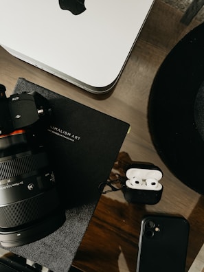 A workspace setup featuring a camera with a large lens, an open case containing wireless earbuds, a smartphone, a notebook titled 'Minimalism Art,' and a Mac computer. The items are arranged on a wooden surface, projecting a sleek and professional aesthetic.