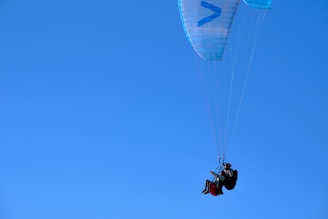 A person is paragliding against a clear blue sky, seated in a harness attached to a parachute-like canopy. The canopy is primarily light blue with a few geometric patterns, and the paraglider is dressed in dark gear, wearing a helmet for safety.