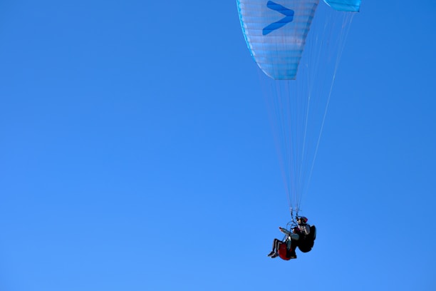 A person is paragliding against a clear blue sky, seated in a harness attached to a parachute-like canopy. The canopy is primarily light blue with a few geometric patterns, and the paraglider is dressed in dark gear, wearing a helmet for safety.