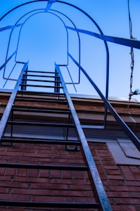 A metal ladder is attached to the side of a red brick building, leading upwards against a clear blue sky. The ladder has protective hoops above it, providing a sense of structure and safety.