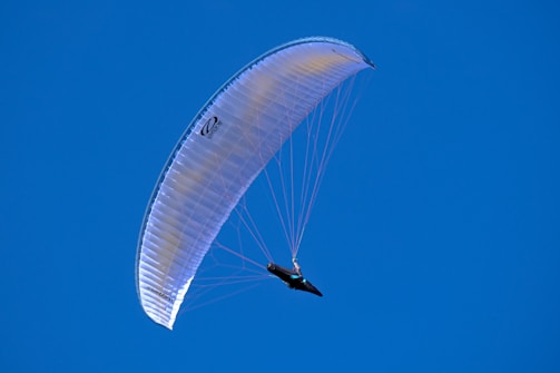 A paraglider soars through the sky under a bright blue canopy. The clear sky provides a vivid backdrop, highlighting the paragliding activity. The glider is suspended beneath the arched wing with strings visible, illustrating the structure and mechanics of paragliding.