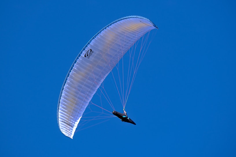 A paraglider soars through the sky under a bright blue canopy. The clear sky provides a vivid backdrop, highlighting the paragliding activity. The glider is suspended beneath the arched wing with strings visible, illustrating the structure and mechanics of paragliding.