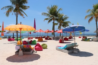 A vibrant beach scene with colorful umbrellas and bean bags scattered across the sandy shore. Several palm trees provide a tropical backdrop, and people relax on the chaise lounges under the shade of the umbrellas. The ocean is visible in the background under a clear blue sky.