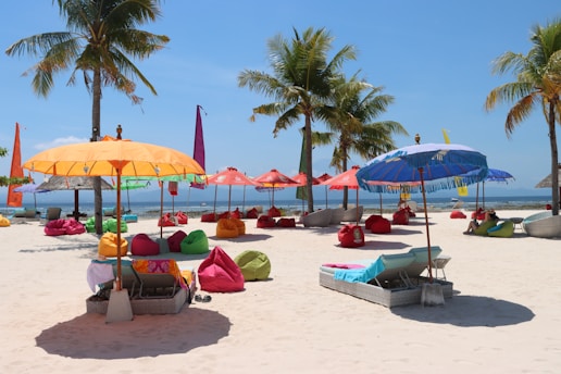 A vibrant beach scene with colorful umbrellas and bean bags scattered across the sandy shore. Several palm trees provide a tropical backdrop, and people relax on the chaise lounges under the shade of the umbrellas. The ocean is visible in the background under a clear blue sky.