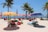 A vibrant beach scene with colorful umbrellas and bean bags scattered across the sandy shore. Several palm trees provide a tropical backdrop, and people relax on the chaise lounges under the shade of the umbrellas. The ocean is visible in the background under a clear blue sky.