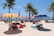 A vibrant beach scene with colorful umbrellas and bean bags scattered across the sandy shore. Several palm trees provide a tropical backdrop, and people relax on the chaise lounges under the shade of the umbrellas. The ocean is visible in the background under a clear blue sky.