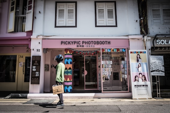A person wearing a helmet, a green jacket, and carrying a brown shopping bag is walking in front of a colorful photobooth shop. The shop is named PickyPic Photobooth and has decorations and photo samples displayed outside. The store is located in a street lined with other buildings and has a modern, vibrant design with shades of pink and multicolored patterns.