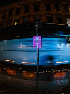 A brightly colored sign encouraging people to dispose of their chewing gum properly is seen in front of a blurred, fast-moving vehicle at night. The background features the facade of an old building with large windows, and the ground is wet, reflecting the vehicle's lights.
