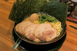 A bowl of ramen featuring rich broth, thick noodles, slices of pork, a boiled egg, and fresh greens, all topped with sheets of seaweed. The dish is arranged in a black bowl resting on a wooden surface with a chopstick holder nearby.