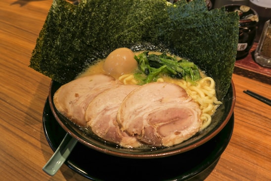 A bowl of ramen featuring rich broth, thick noodles, slices of pork, a boiled egg, and fresh greens, all topped with sheets of seaweed. The dish is arranged in a black bowl resting on a wooden surface with a chopstick holder nearby.
