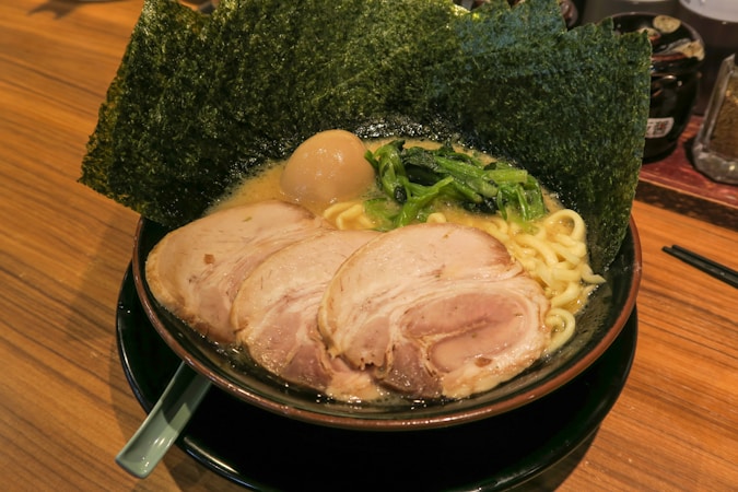 A bowl of ramen featuring rich broth, thick noodles, slices of pork, a boiled egg, and fresh greens, all topped with sheets of seaweed. The dish is arranged in a black bowl resting on a wooden surface with a chopstick holder nearby.