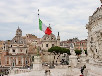 A cityscape featuring historical European architecture with two prominent domed buildings. An Italian flag is prominently displayed on a flagpole in the foreground. The sky is overcast, and several statues and sculptures are visible on a nearby building. A group of manicured trees stands to the right of the domed structures, and a number of people can be seen walking around the area.